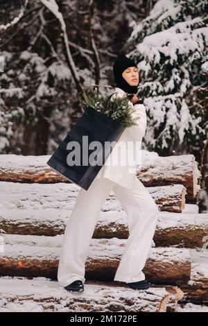 Ein Mädchen in einem weißen Anzug und Balaklava mit einem Paket Weihnachtsbäume im Winterwald an Silvester. Neujahrskonzept Stockfoto