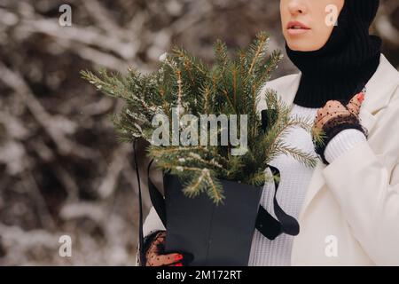 Ein Mädchen in einem weißen Anzug und Balaklava mit einem Paket Weihnachtsbäume im Winterwald an Silvester. Neujahrskonzept Stockfoto