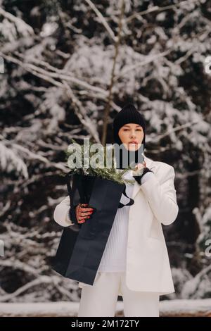 Ein Mädchen in einem weißen Anzug und Balaklava mit einem Paket Weihnachtsbäume im Winterwald an Silvester. Neujahrskonzept Stockfoto