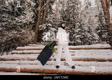 Ein Mädchen in einem weißen Anzug und Balaklava mit einem Paket Weihnachtsbäume im Winterwald an Silvester. Neujahrskonzept Stockfoto