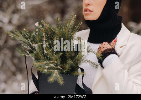Ein Mädchen in einem weißen Anzug und Balaklava mit einem Paket Weihnachtsbäume im Winterwald an Silvester. Neujahrskonzept Stockfoto