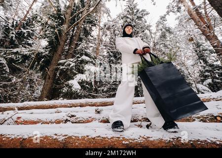 Ein Mädchen in einem weißen Anzug und Balaklava mit einem Paket Weihnachtsbäume im Winterwald an Silvester. Neujahrskonzept Stockfoto
