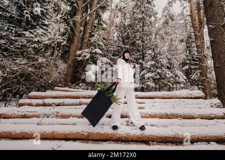 Ein Mädchen in einem weißen Anzug und Balaklava mit einem Paket Weihnachtsbäume im Winterwald an Silvester. Neujahrskonzept Stockfoto