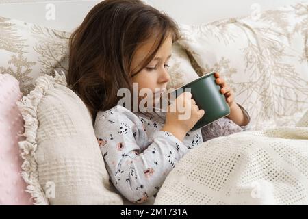 Süßes Kind, kleines dunkelhaariges Vorschulmädchen, trinkt heiße, ungenießbare Medizin aus grüner Tasse, liegt im Pyjama in einem weichen Bett. Genesung, Gesundheitsfürsorge. Stockfoto