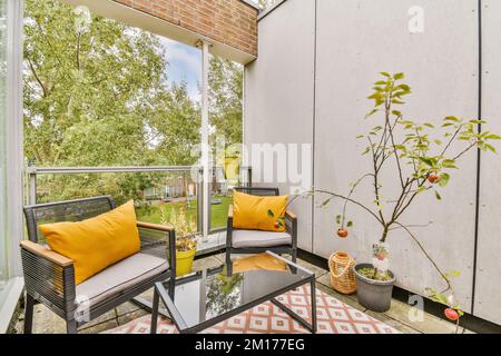 Eine Terrasse mit zwei Stühlen und einem Apfelbaum auf der Rückseite, auf einem Glastisch umgeben von orangefarbenen Kissen Stockfoto