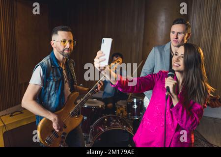 Mittellange Aufnahme einer Musikband junger Leute, die ein Selfie bei Wiederholungen im Studio macht, Musikkonzept. Hochwertiges Foto Stockfoto