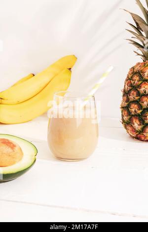 Bananen-, Ananas- und Avocado-Smoothie auf einem weißen Holztisch bei Sonnenlicht. Minimal-Entgiftungsdiät-Konzept, Sommer-Vitamingetränk. Stockfoto