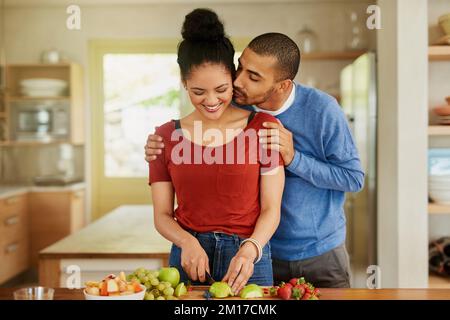Sich gegenseitig dabei zu unterstützen, die gesündesten zu sein, die sie sein können. Aufnahme eines jungen Mannes, der seine Frau küsst, während sie zu Hause einen gesunden Snack zubereitet. Stockfoto