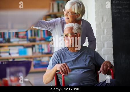Nichts bringt uns in den Boden. Aufnahme einer älteren Frau, die ihren Mann zu Hause in einen Rollstuhl schiebt. Stockfoto