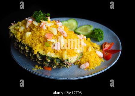 Gebratener Reis mit Meeresfrüchten in einer Ananas mit Gemüse auf blauem Teller, isoliert auf schwarzem Hintergrund, Draufsicht Stockfoto