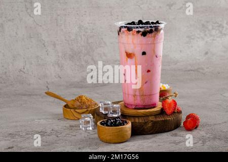 Boba- oder Tapioka-Perlen sind taiwanischer Seifenmilchtee in Plastikbecher mit Erdbeermarmelade auf texturiertem Hintergrund, Sommererfrischung. Stockfoto