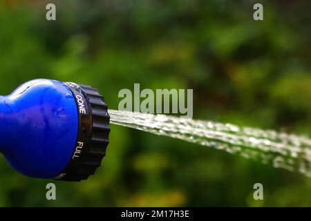 Unschärfe-Effekte im Wassergarten. Eine weibliche Hand, die blaue automatische Sprinkler hält, sprüht Wasser auf braunen, unscharfen Hintergrund. Gärtner, der den Rasen bewässert Stockfoto