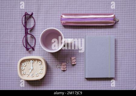 Schreibwaren, Bürobedarf. Notizbuch, Clip, Becher, Uhr, Brille, Federmäppchen auf einem Stoffhintergrund mit violettem Karomuster. Flach liegend, Draufsicht Stockfoto