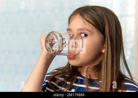 Ein kleines Mädchen trinkt Wasser aus einem Glas Tasse. Kleines Mädchen hält ein Glas Wasser. Durst nach Wasser Stockfoto