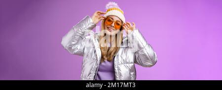 Stilvolle freche gewagte blonde europäische Mädchen handeln cool tragen stilvoll Sonnenbrille Silber Jacke Winter Hut Überprüfung Rahmen Nase kippbaren Kopf Frech lächelnd Stockfoto