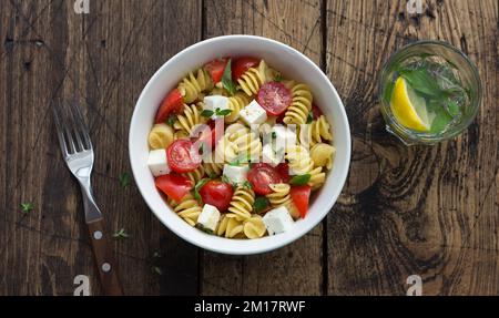 Hausgemachter Salat mit Pasta, Tomaten, Fetakäse und Gewürzen auf einem Holztisch. Einfaches, köstliches Essen. Draufsicht, Platz Stockfoto