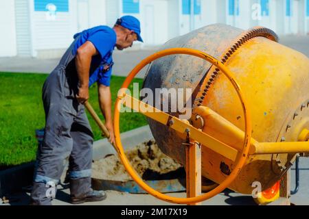 Uniformierter Bauarbeiter arbeitet am Sommertag auf der Baustelle. Alte Maurerschaufel schaufelt Zement und Sand in Betonmischer. Auftragnehmer für die Reparatur von Gebäuden und Strukturen. Stockfoto