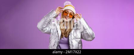 Stilvolle freche gewagte blonde europäische Mädchen handeln cool tragen stilvoll Sonnenbrille Silber Jacke Winter Hut Überprüfung Rahmen Nase kippbaren Kopf Frech lächelnd Stockfoto