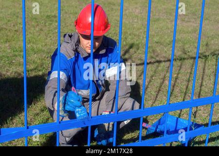 Professioneller Maler in Kleidung und Helm lackiert Metallzaun oder Zaun aus Armierung mit Pinsel am Sommertag. Authentischer Workflow. Echtes Arbeitsfoto... Stockfoto