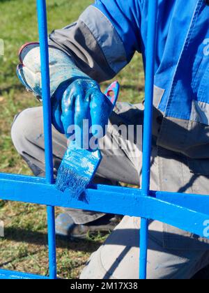 Industrielackierer lackieren Metallzäune oder Zäune blau. Hand des Arbeiters im Overall mit Nahpinsel. Authentischer Workflow. Hintergrund... Stockfoto