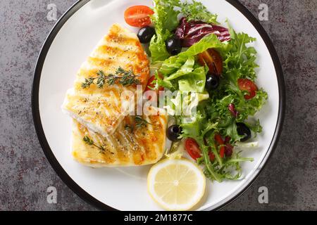 Diät-Food gegrillter Kabeljau mit frischem Salat aus Tomaten, Oliven, Rucola, Salatmischung und Zitrone in Nahaufnahme auf einem Teller auf dem Tisch. Horizontale Draufsicht Stockfoto