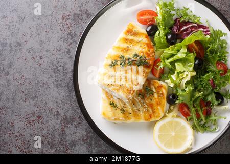 Köstlicher, gesunder gegrillter Kabeljau mit frischem Salat aus Tomaten, Oliven, Salatmischung und Zitrone in Nahaufnahme auf einem Teller auf dem Tisch. Horizontale Draufsicht f Stockfoto