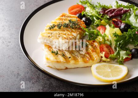 Diät-Food gegrillter Kabeljau mit frischem Salat aus Tomaten, Oliven, Rucola, Salatmischung und Zitrone in Nahaufnahme auf einem Teller auf dem Tisch. Horizontal Stockfoto