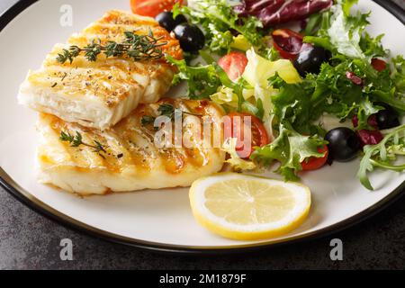 Köstlicher, gesunder gegrillter Kabeljau mit frischem Salat aus Tomaten, Oliven, Salatmischung und Zitrone in Nahaufnahme auf einem Teller auf dem Tisch. Horizontal Stockfoto