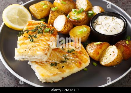 Portion gegrillten Kabeljaus mit gebackenen Jackenkartoffeln, cremiger Sauce und Zitrone in Nahaufnahme auf einem Teller auf dem Tisch. Horizontal Stockfoto