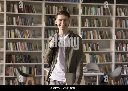 Glücklicher, gutaussehender Teenager-Schüler mit Rucksack, der in der Bibliothek steht Stockfoto