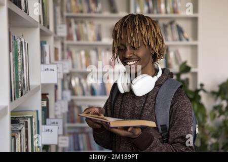 Glücklicher afrikanischer Student in der Universitätsbibliothek, steht an Bücherregalen Stockfoto