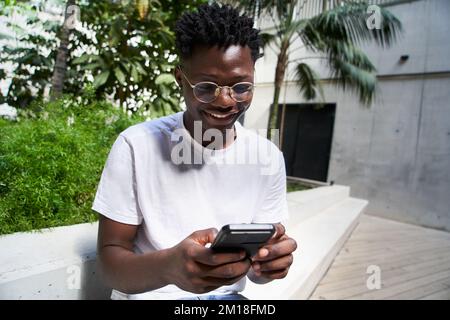 Porträt eines glücklichen Schwarzen, der ein Handy benutzt und auf dem Universitätscampus lacht. Stockfoto