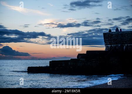 Brighton, Dezember 10. 2022: Besucher, die von einem der Groynes aus auf das Meer blicken, während die Sonne am Brighton Beach untergeht Stockfoto