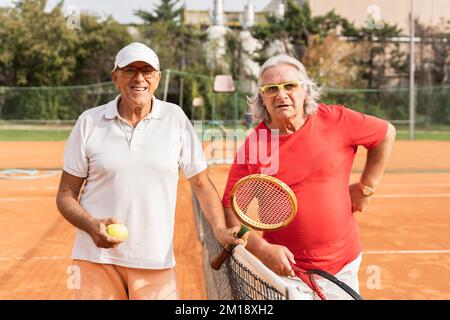 Porträt von zwei älteren Tennisspielern in Sportbekleidung, die auf einem Sandtennisplatz die Hände schütteln - Wellnesskonzept für Rentner Stockfoto