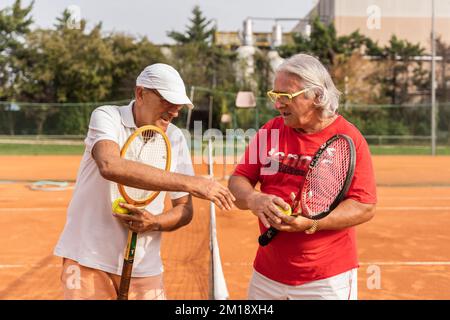 Porträt von zwei älteren Tennisspielern in Sportbekleidung, die auf einem Sandtennisplatz die Hände schütteln - Wellnesskonzept für Rentner Stockfoto