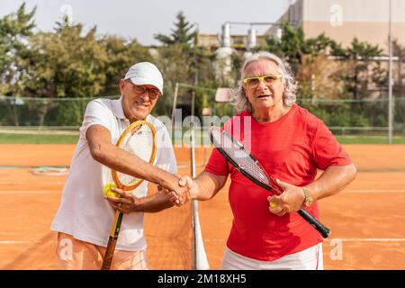 Porträt von zwei älteren Tennisspielern in Sportbekleidung, die auf einem Sandtennisplatz die Hände schütteln - Wellnesskonzept für Rentner Stockfoto