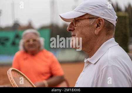 Porträt von zwei älteren Tennisspielern in Sportbekleidung, die sich am Ende des Spiels auf einem Sandtennisplatz entspannen - Wellnesskonzept für Rentner Stockfoto