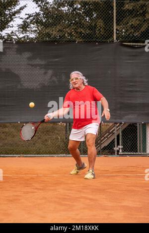 Senior-Tennisspieler in Sportbekleidung in Aktion auf einem Sandtennisplatz - Wellness-Konzept für Rentner Stockfoto