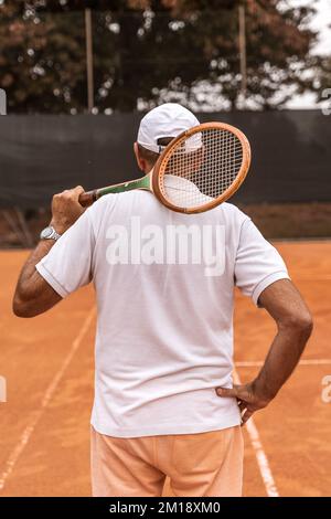 Ein pensionierter Tennisspieler in Sportbekleidung posiert mit dem Rücken zur Kamera auf einem Sandtennisplatz - Wellness-Konzept für Rentner Stockfoto