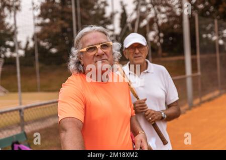 Porträt von zwei älteren Tennisspielern in Sportbekleidung, die sich am Ende des Spiels auf einem Sandtennisplatz entspannen - Wellnesskonzept für Rentner Stockfoto