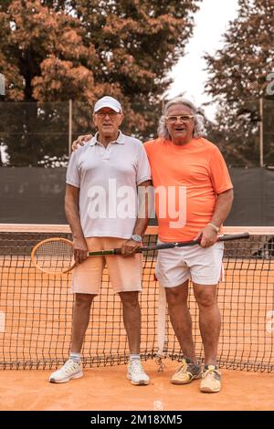 Porträt von zwei älteren Tennisspielern in Sportbekleidung, die am Ende des Spiels auf einem Sandtennisplatz posieren - Wellness-Konzept für Rentner Stockfoto