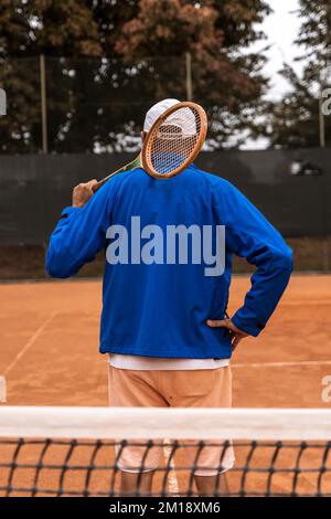 Ein pensionierter Tennisspieler in Sportbekleidung posiert mit dem Rücken zur Kamera auf einem Sandtennisplatz - Wellness-Konzept für Rentner Stockfoto