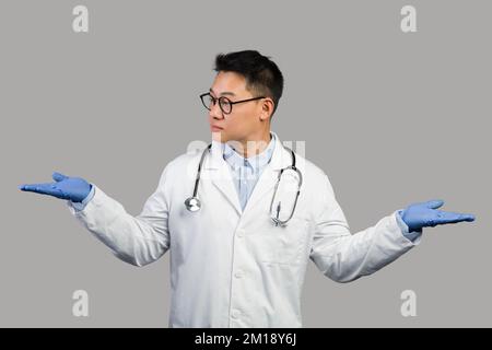 Seriöser, reifer chinesischer Therapeut in weißem Mantel und Brille spreizt seine Arme an die Seiten und hält leeren Raum Stockfoto
