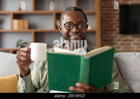 Ein glücklicher, reifer Schwarzer mit Brille genießt eine Tasse Kaffee und liest in der Freizeit im Wohnzimmer, aus nächster Nähe Stockfoto