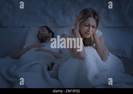 Depressive Frau, die unter Schnarchen leidet, schlafender Ehemann, Frau, die die Ohren mit Händen bedeckt, die nachts auf dem Bett sitzt Stockfoto