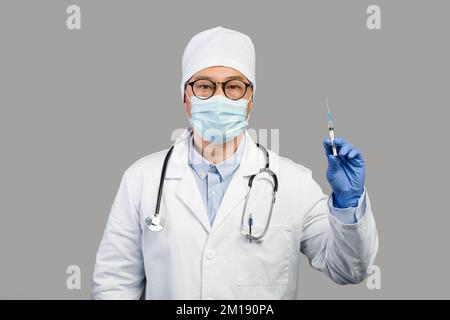 Seriöser reifer chinesischer Mann Therapeut in weißem Mantel und Schutzmaske zeigt Spritze mit Impfstoff Stockfoto