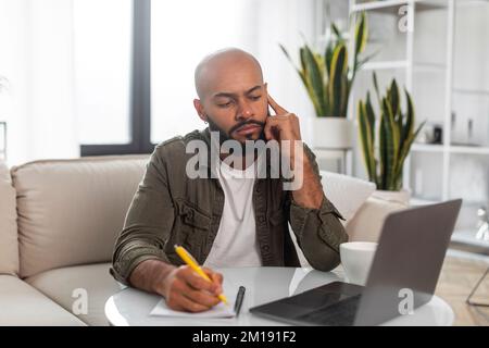 Ein ernster Mann, der sich am Handy unterhält und Notizen macht, auf den Laptop-Bildschirm schaut, zu Hause auf dem Sofa sitzt Stockfoto