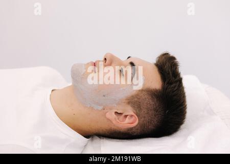 Kosmetologe mit Tonmaske auf das Gesicht des Mannes im Wellnesssalon. Seitenansicht des männlichen Gesichts während des Eingriffs. Stockfoto