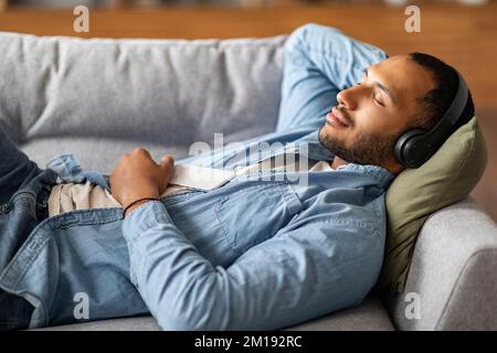 Junger Schwarzer Mit Kabellosen Kopfhörern, Der Sich Auf Dem Sofa Mit Dem Smartphone Entspannen Kann Stockfoto