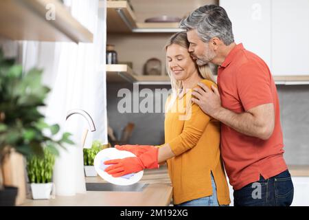 Attraktive reife blonde Frau, die Geschirr spült, Ehemann küsst sie Stockfoto